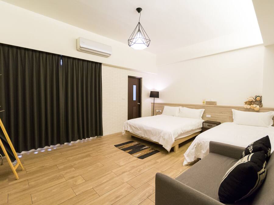A hotel room with two beds, a sofa, a wooden floor, a black curtain, a lamp, and a hanging light fixture.
