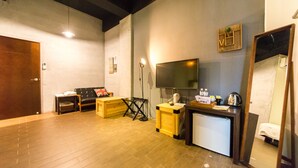 Habitación de hotel con piso de madera, un escritorio con computadora, un televisor, un espejo y un pequeño rincón de descanso.