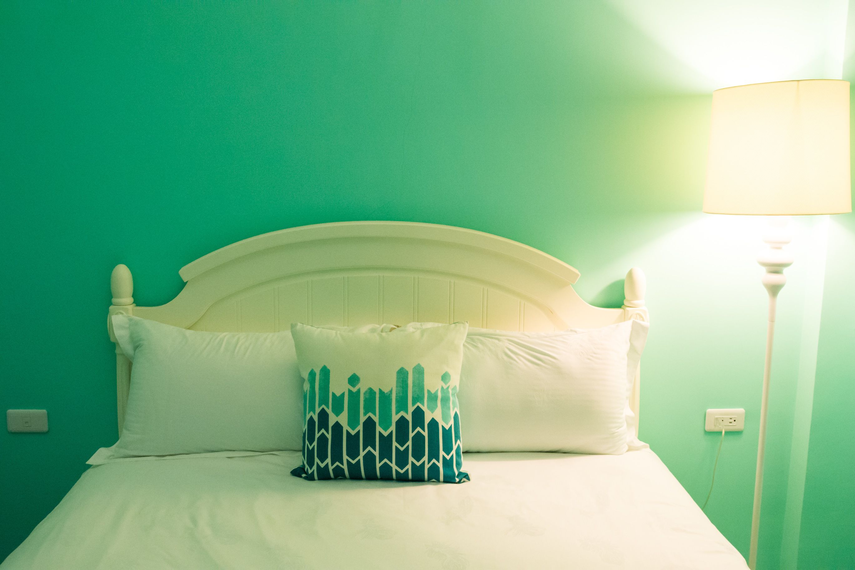 A bed with white bedding and a patterned pillow, a white headboard, and a lamp on a green wall.