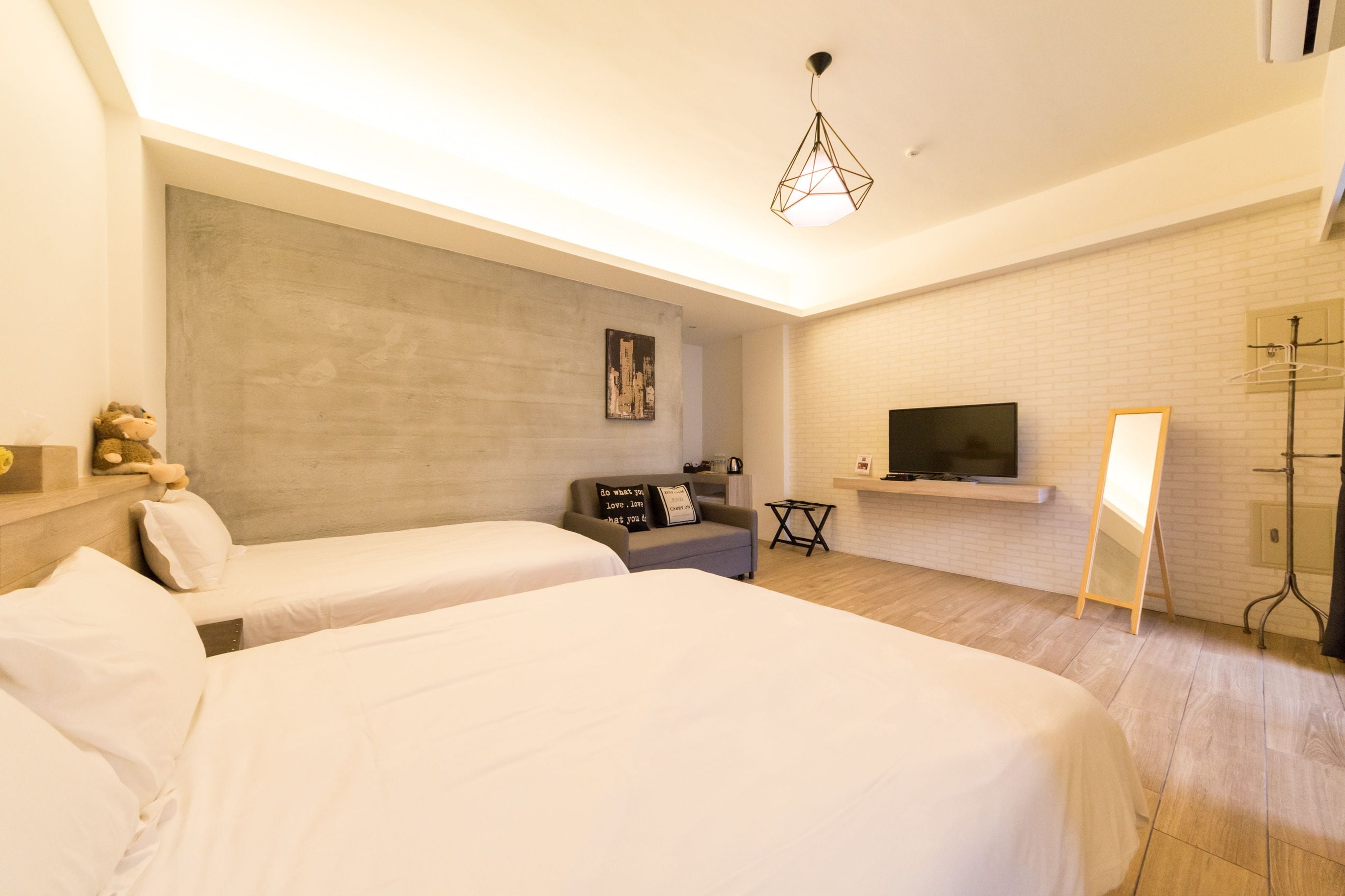 Quarto de hotel com duas camas, um sofá cinzento, uma TV presa a uma parede de tijolos e um candeeiro suspenso.