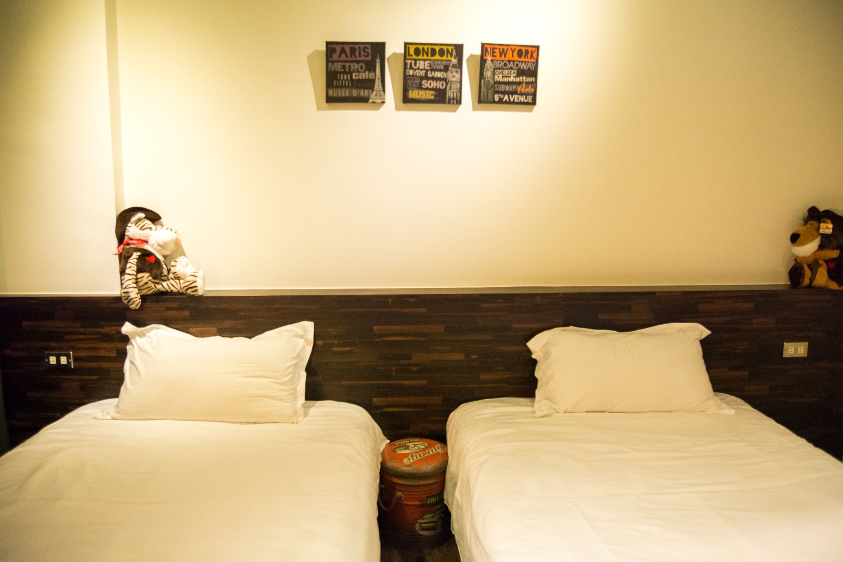 A hotel room with two beds, a wooden headboard, and three signs on the wall advertising Paris, London, and New York.