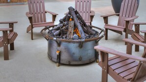 Uma lareira de metal com lenha, rodeada por cadeiras Adirondack de madeira.