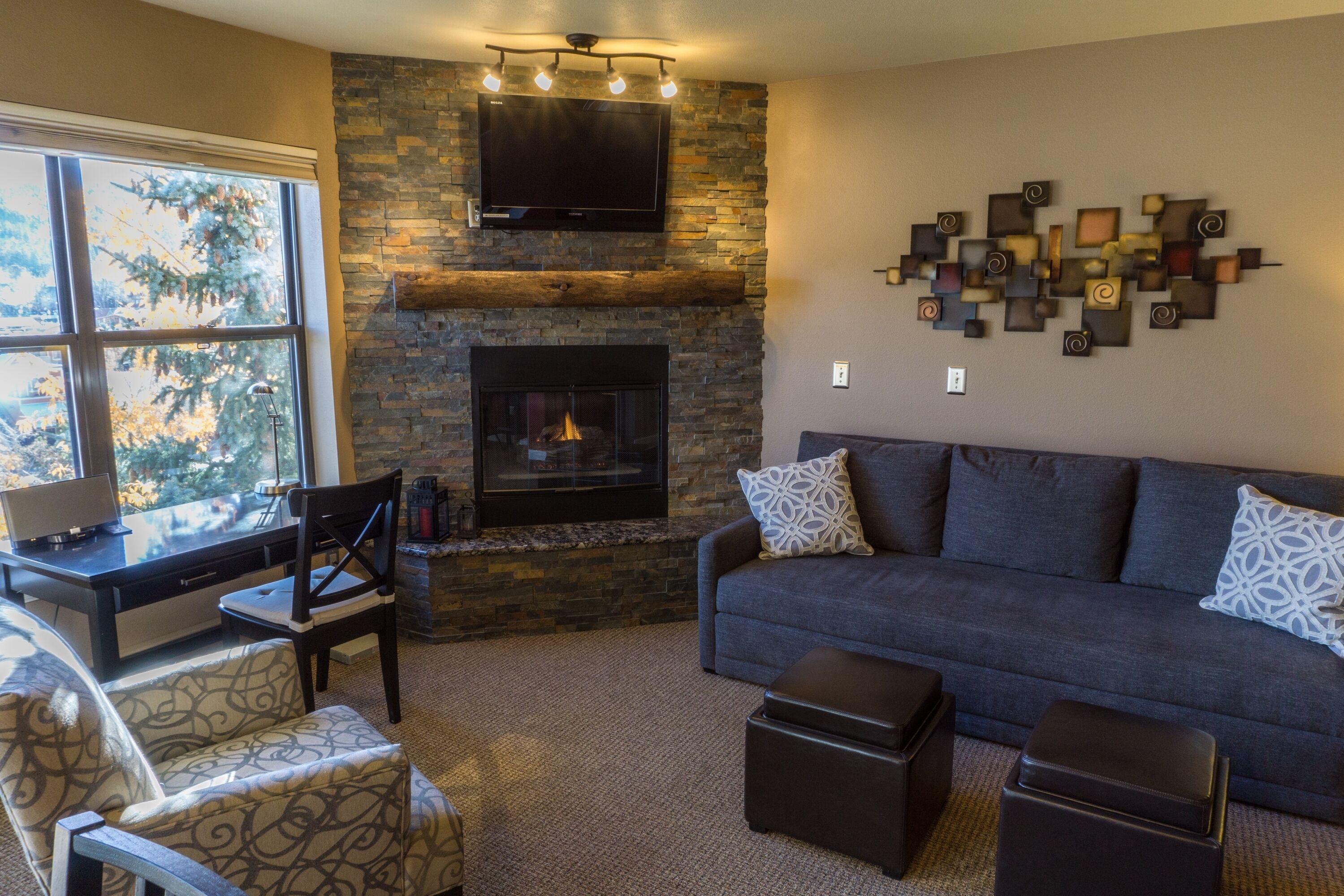 A living room with a stone fireplace, a gray sofa, patterned chairs, and a flat-screen TV mounted above the fireplace.
