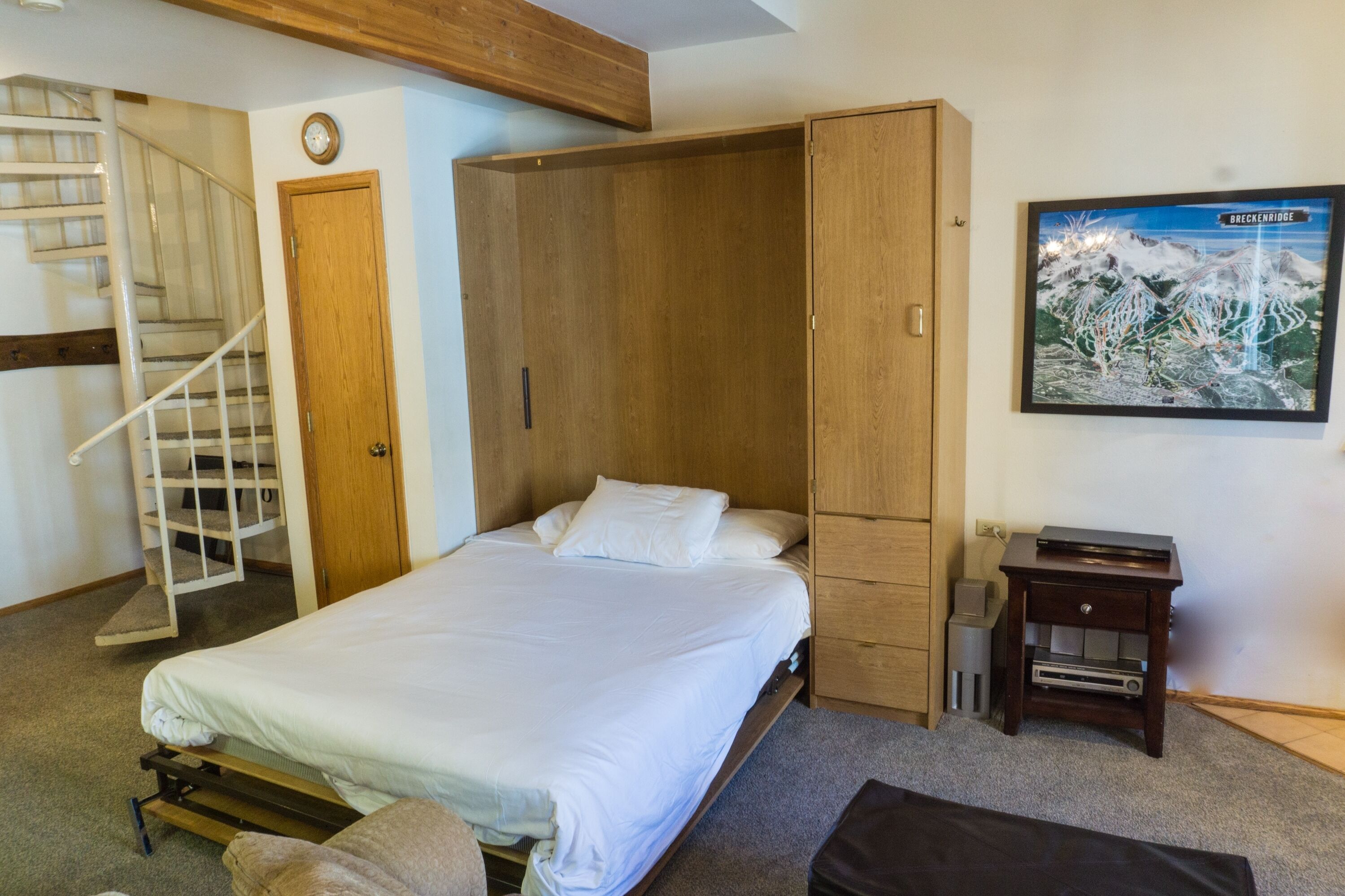 Un dormitorio con cama, un armario de madera, una mesita de noche pequeña de madera, un televisor montado en la pared y una escalera.