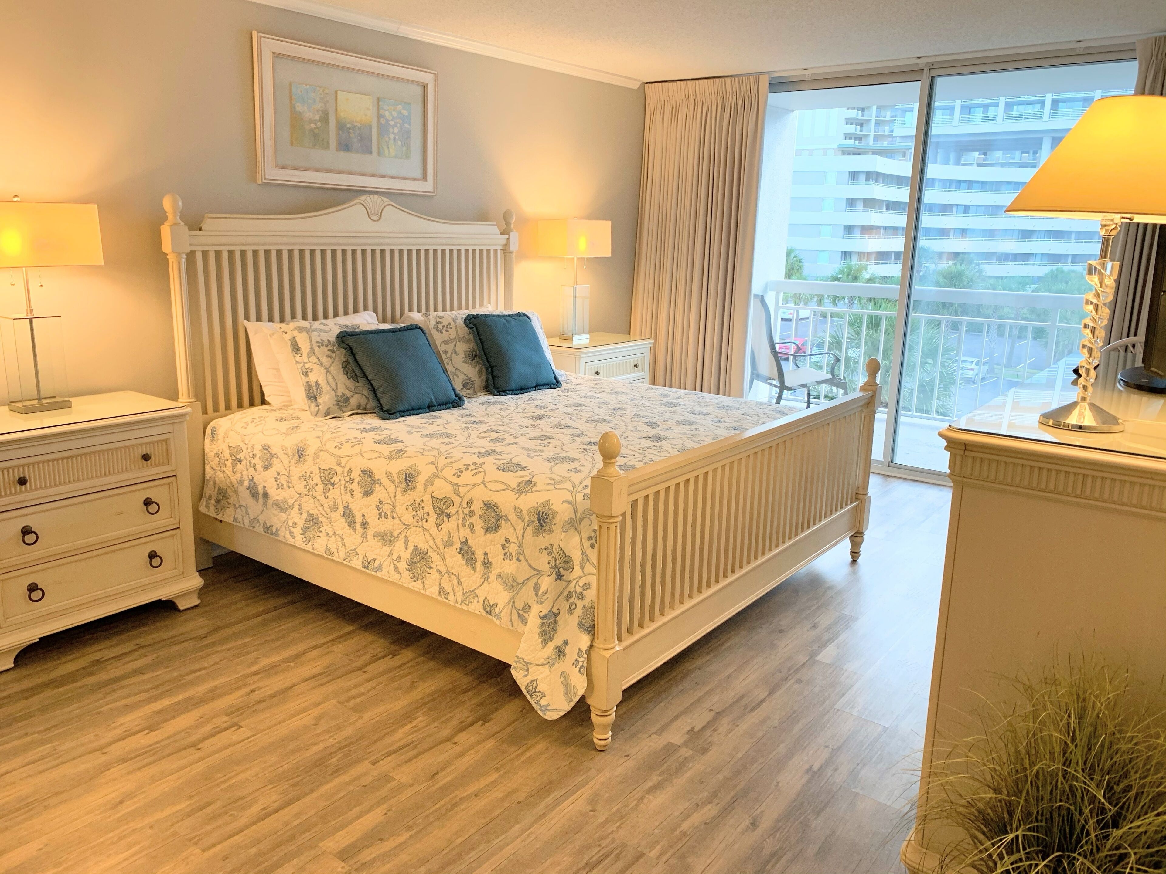 A bedroom with a large bed, two bedside tables, a nightstand, a dresser, a window with curtains, and a view of buildings.