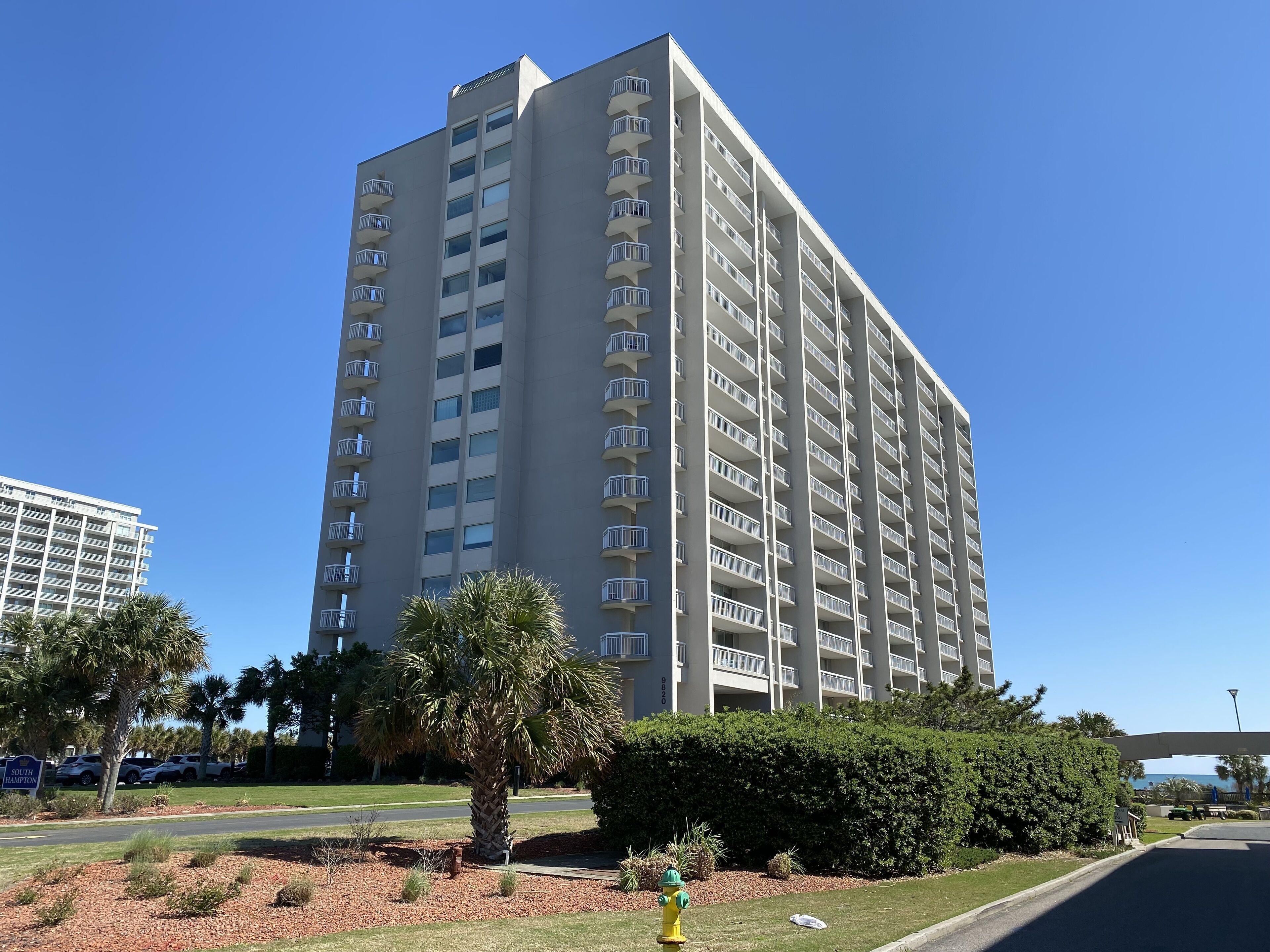 A tall, multi-story building with balconies, surrounded by palm trees and a well-maintained lawn.
