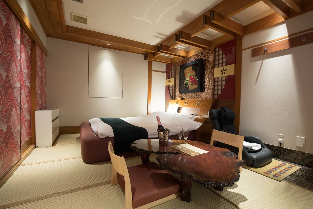 A traditional Japanese-style room with tatami flooring, a bed with a white and black bedspread, a wooden table with a bottle of champagne, and a massage chair.