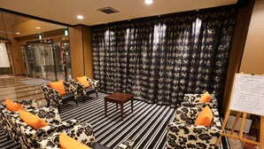 Een hotellobby met een zwart-wit gestreept tapijt, banken met bloemenmotief en oranje kussens, een klein houten tafeltje en een bordje op een standaard.