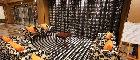Un hall d’hôtel avec un tapis à rayures noires et blanches, des canapés à motifs floraux ornés de coussins orange, une petite table en bois et un panneau d’affichage sur un présentoir.