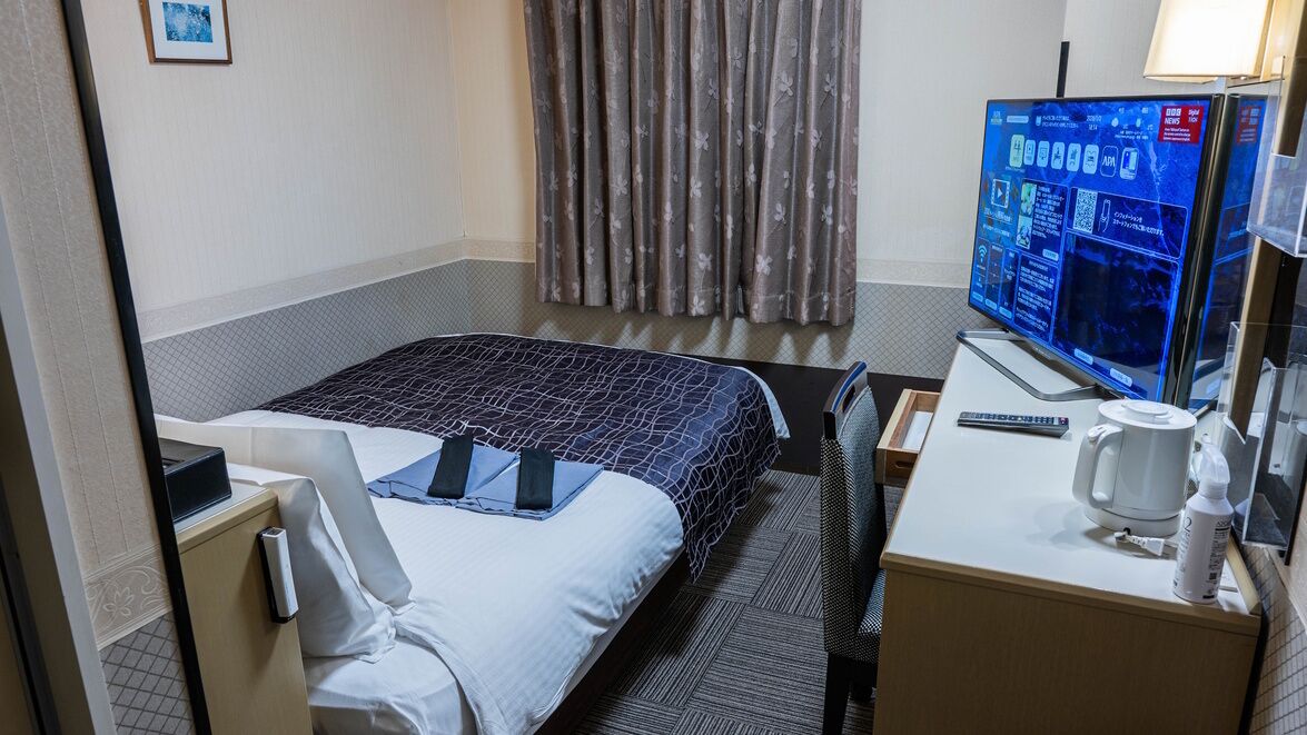 Une chambre d’hôtel avec un lit, un bureau doté d’un écran d’ordinateur, une machine à café et une fenêtre avec des rideaux.
