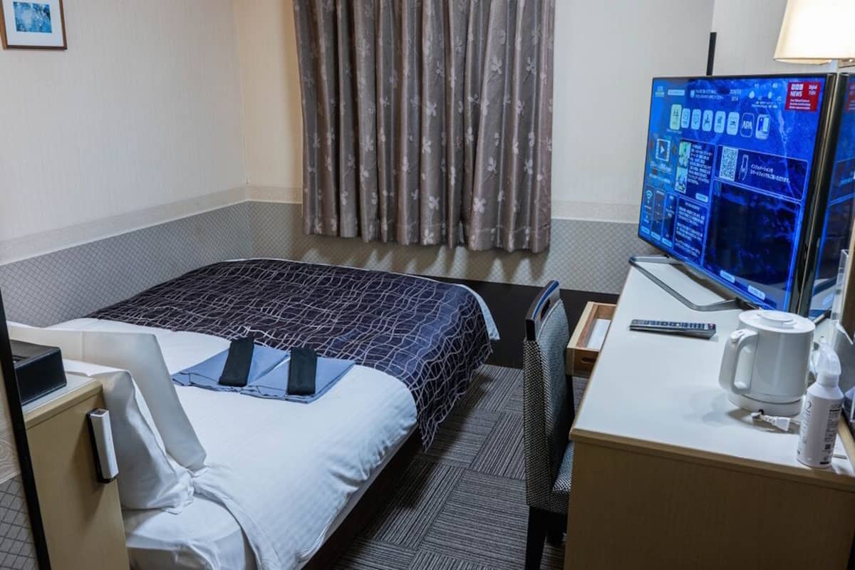 Une chambre d’hôtel avec un lit, un bureau doté d’un écran d’ordinateur, une machine à café et une fenêtre avec des rideaux.