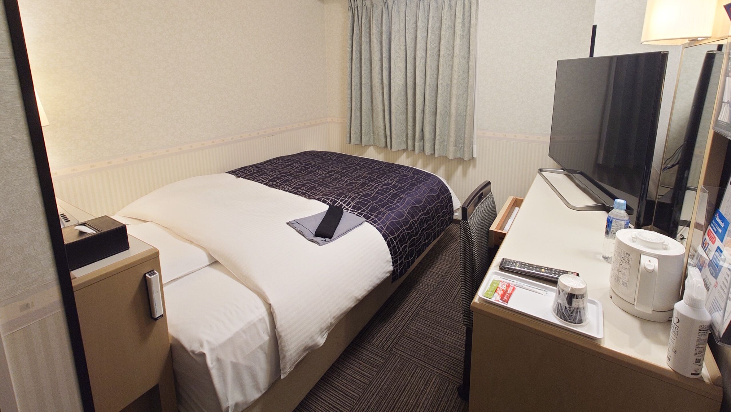 Ein Hotelzimmer mit einem Bett, einem Schreibtisch mit Fernseher, einem Sessel, einem Nachttisch und einem Fenster mit Vorhängen.