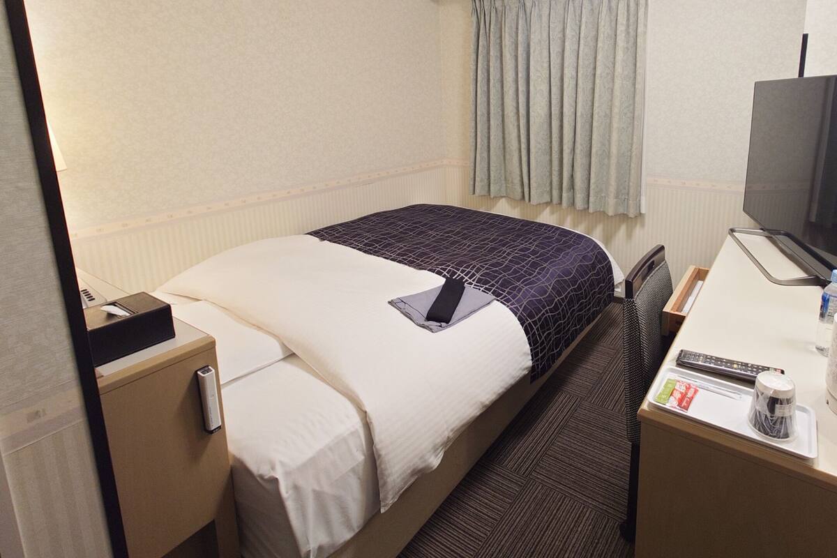Une chambre d’hôtel comprenant un lit, un bureau avec une télévision, une chaise, une table de chevet et une fenêtre avec des rideaux.