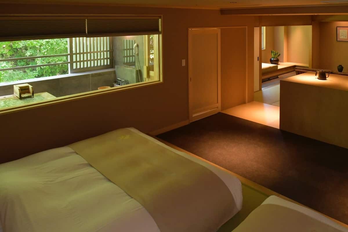 A hotel room with a bed, a small table, a window with a view of greenery, and a bathroom with a shower.