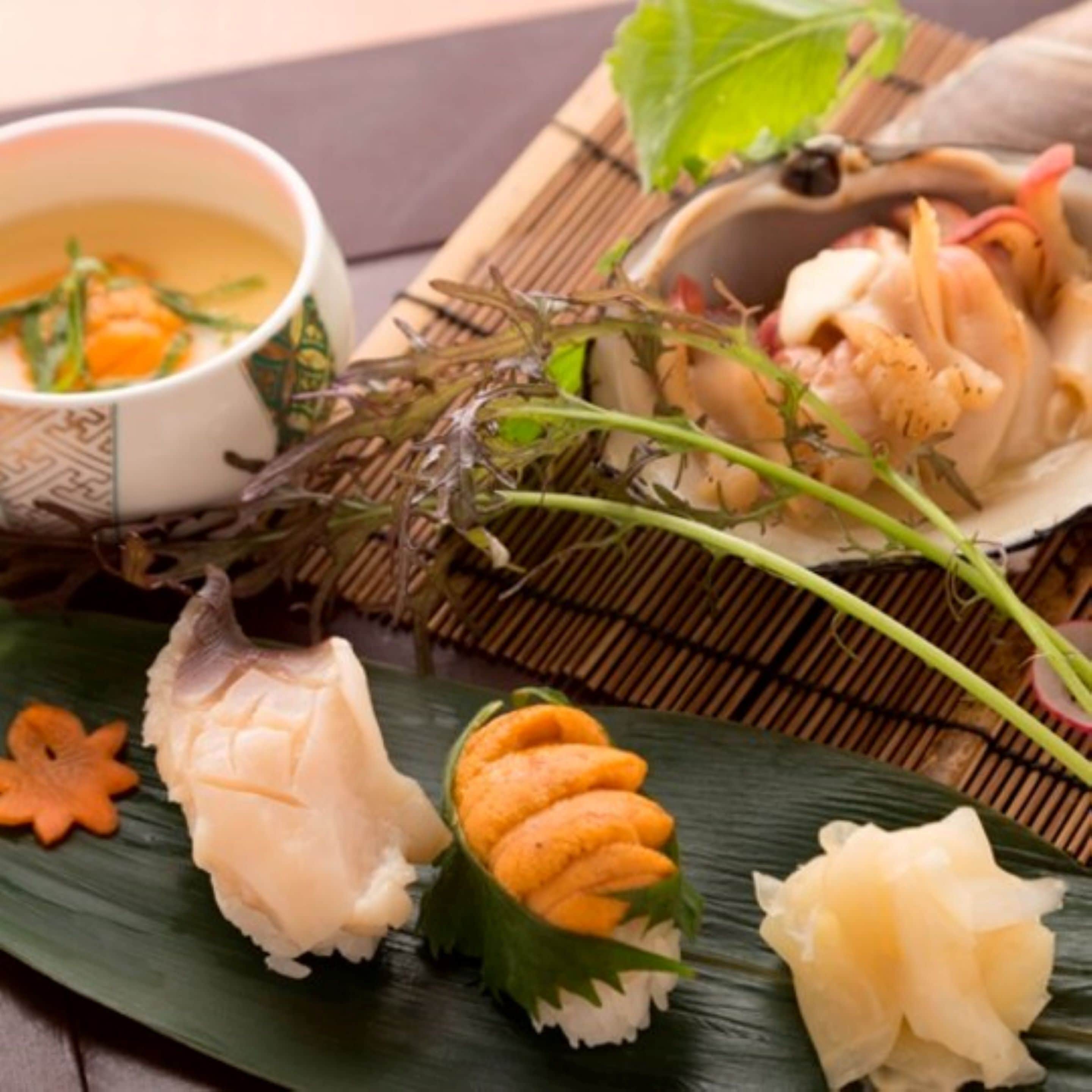 Variedad de sushi y sashimi en un plato sobre hojas verdes, acompañado de un tazón de sopa y un recipiente pequeño con mariscos.