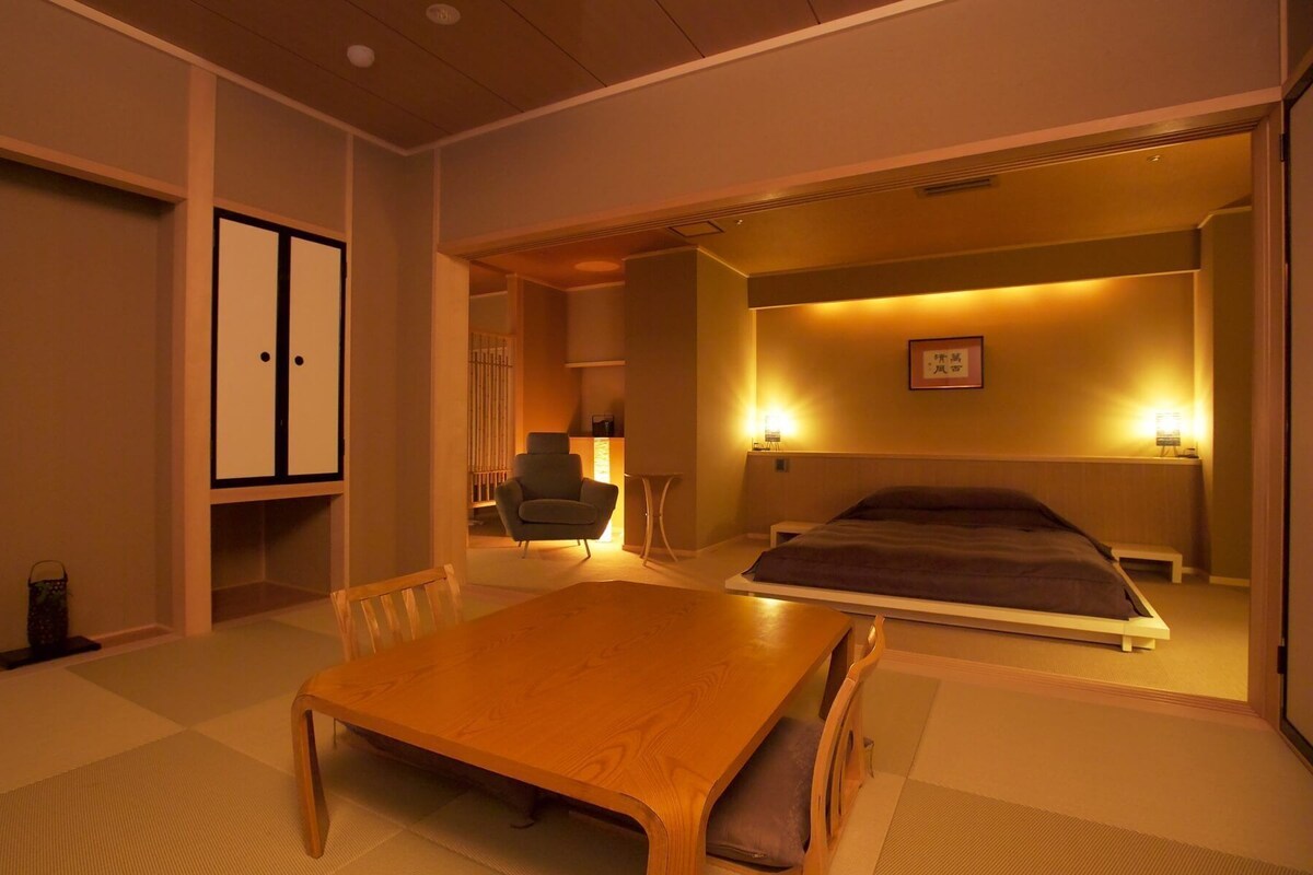 Une pièce de style japonais traditionnel, avec un sol recouvert de tatamis, une petite table en bois, un lit avec une couverture sombre et un petit coin salon avec une chaise.