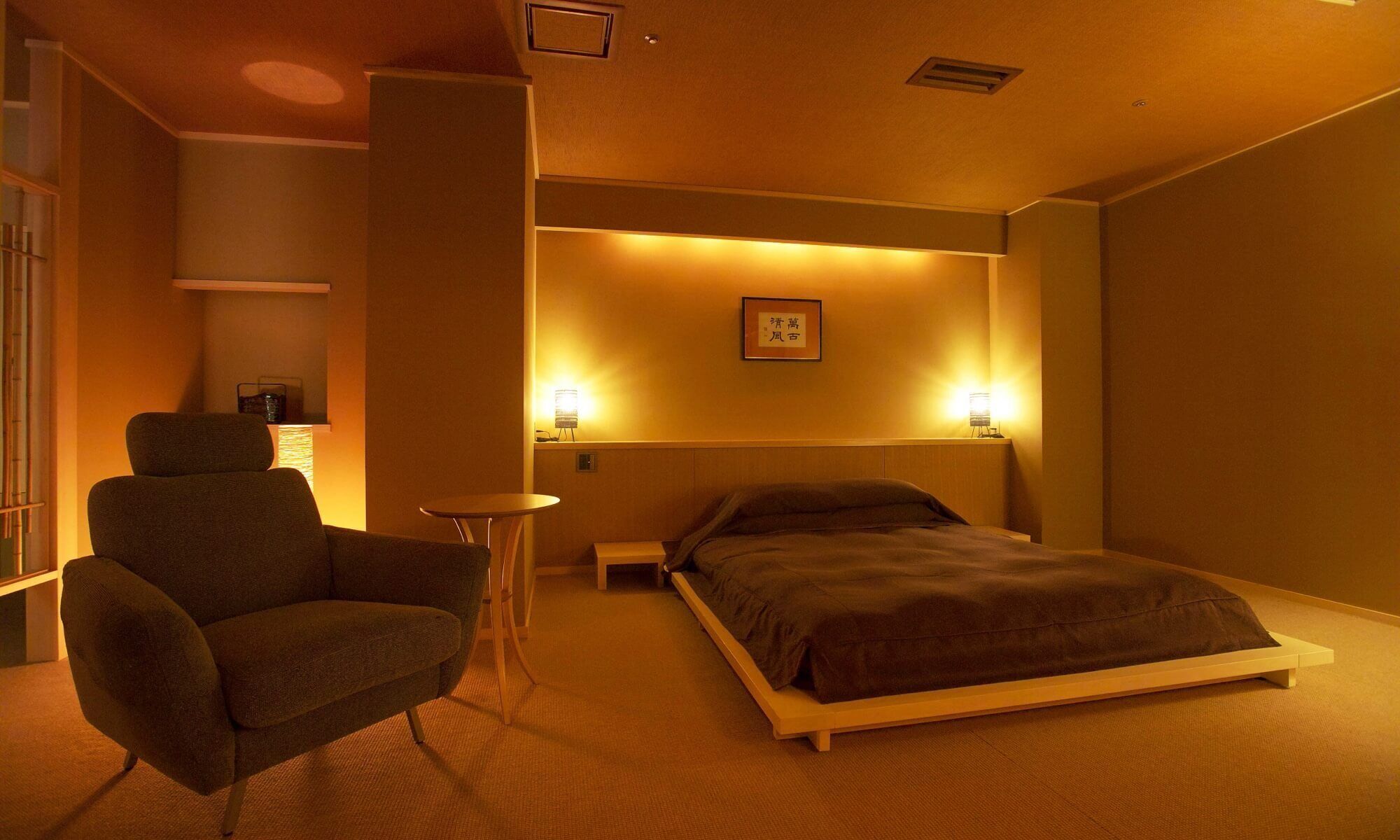 Een hotelkamer met een bed, een stoel, een klein tafeltje en twee wandlampen.