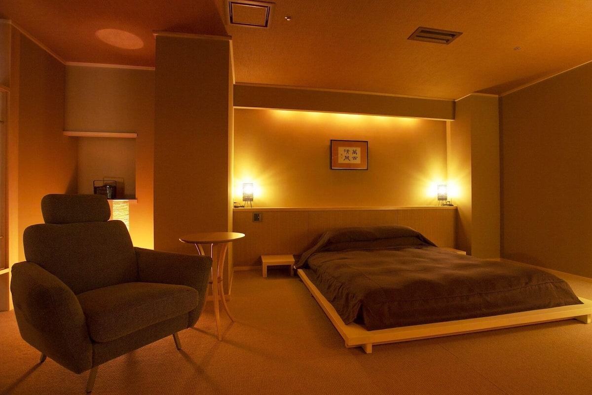 A hotel room with a bed, a chair, a small table, and two wall-mounted lamps.