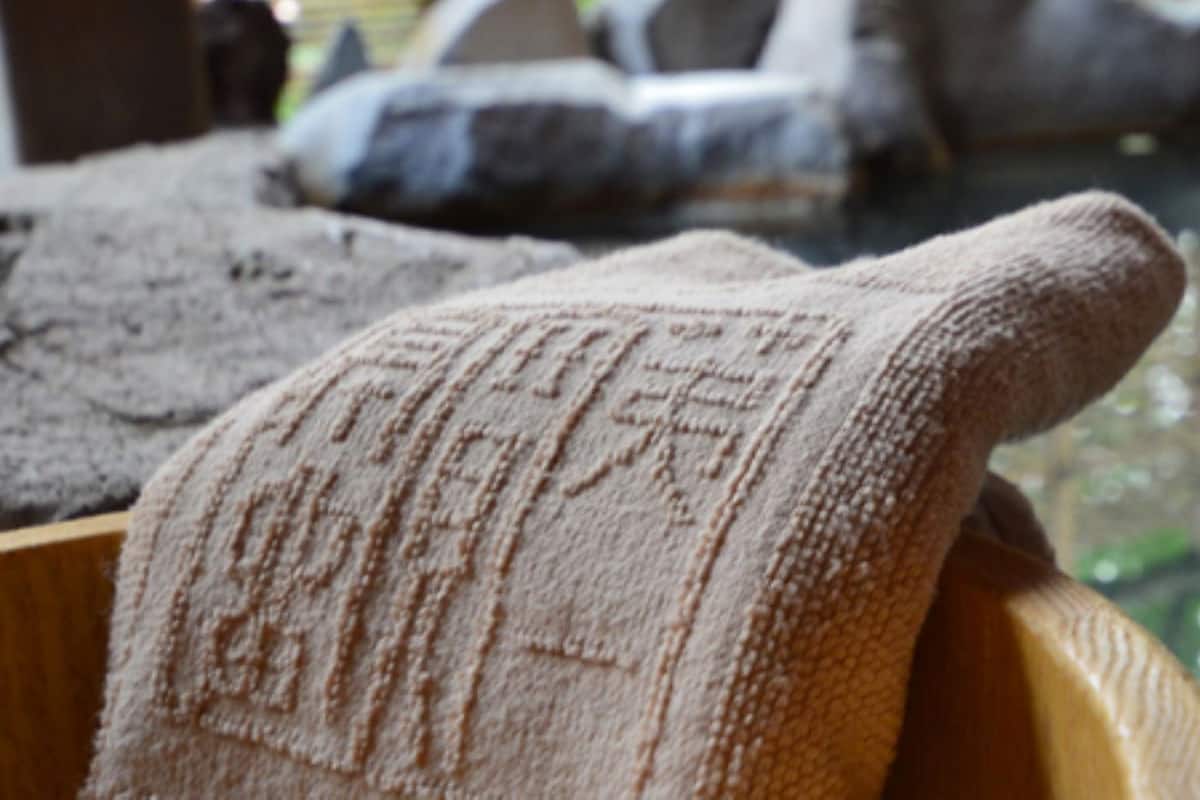 A wooden bowl with a towel draped over it, placed near a body of water, possibly a pond or small lake, with rocks and a wooden structure in the background.