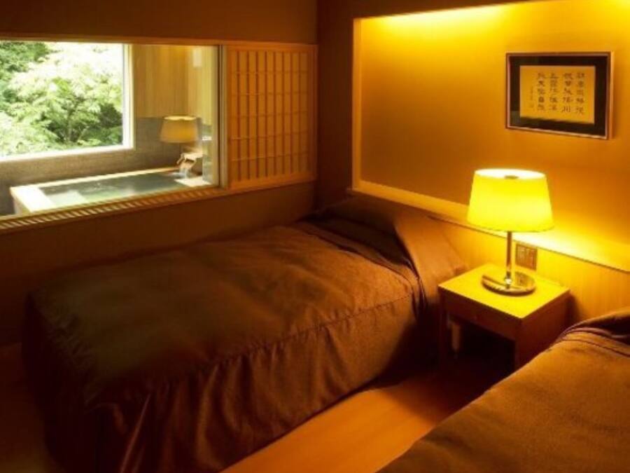 A hotel room with two beds, a window with a view of greenery, a bedside table with a lamp, and a framed picture on the wall.