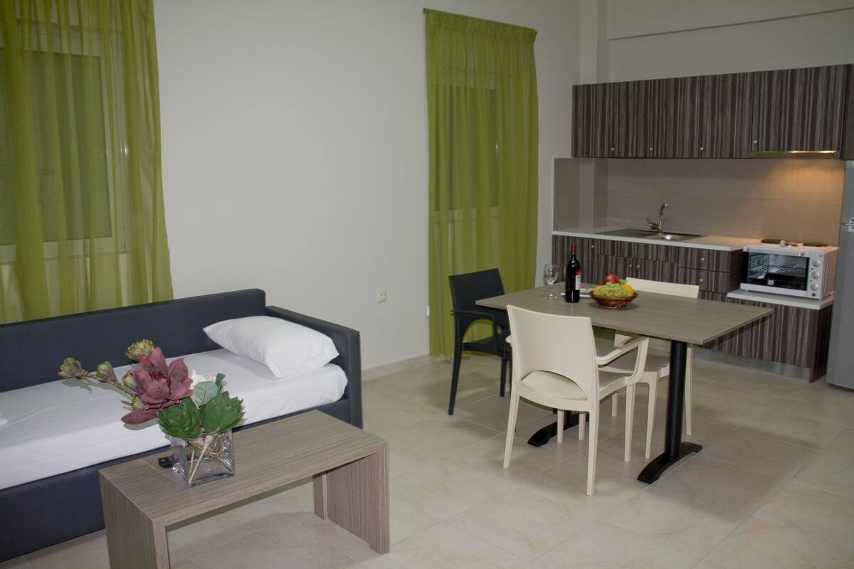 A modern hotel room with a bed, a small dining table, and a kitchenette.