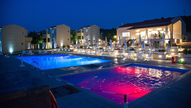 A pool area with multiple pools and lounge chairs, surrounded by buildings with outdoor seating and a covered area. - Fournia Village (Andravida-Kyllini)