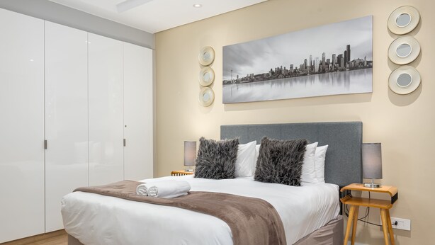 A modern bedroom with a large bed, a cityscape painting on the wall, and a bedside table with a lamp. - Warwick 504 (Cape Town)