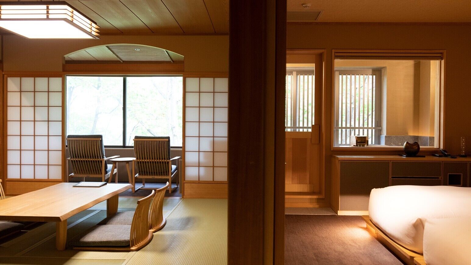 Una habitación tradicional japonesa con suelo de tatami, una mesa baja, sillas, una cama y puertas correderas.