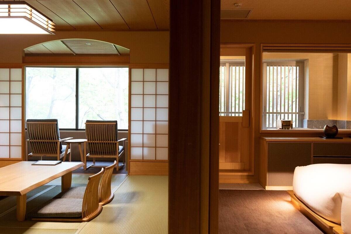 Quarto japonês tradicional com tatames, uma mesa baixa, cadeiras, uma cama e portas deslizantes.