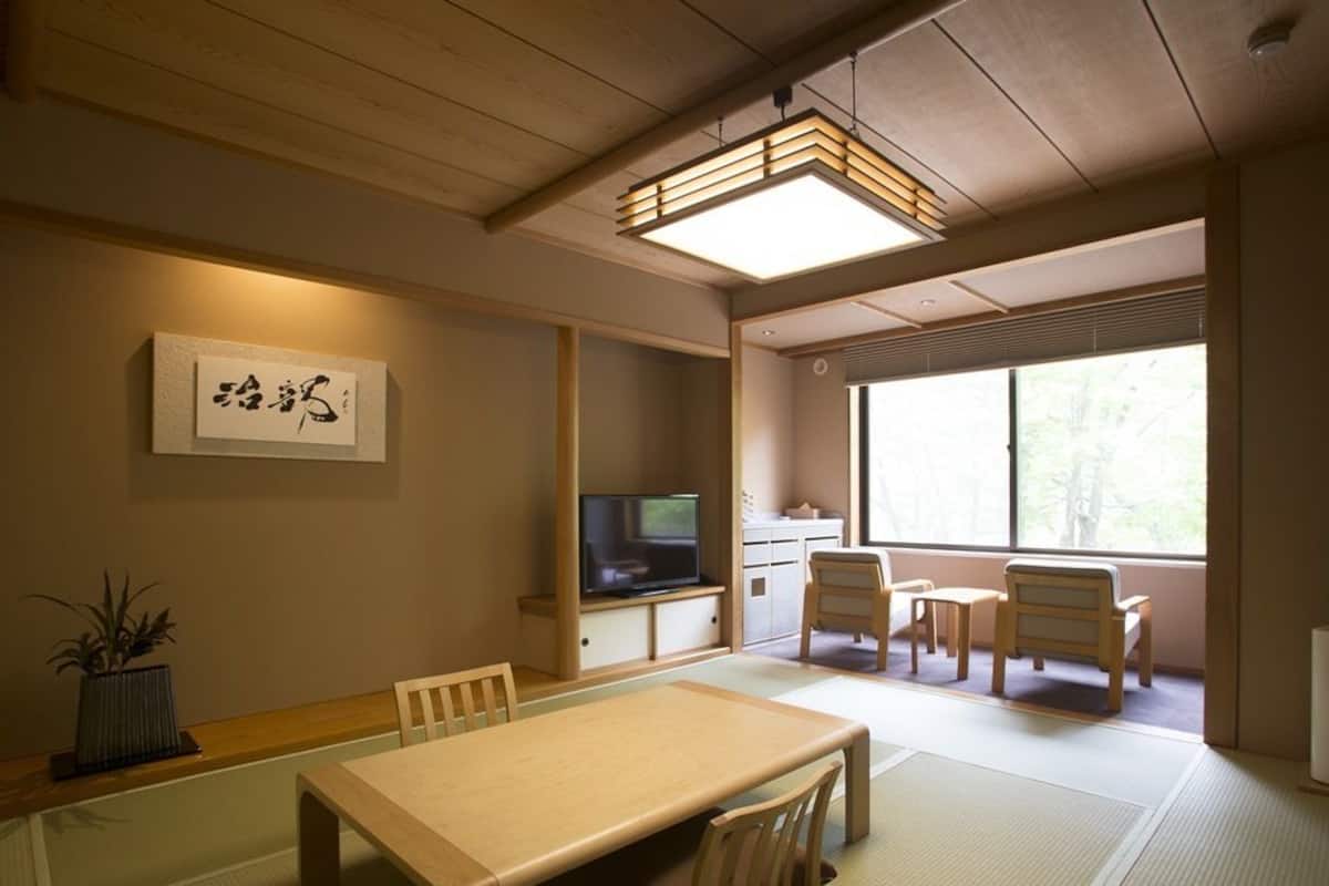 Quarto japonês tradicional com tatames, uma mesa baixa, cadeiras, televisão e janela com vista para árvores.