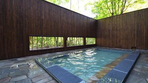 A spa pool with stone flooring and wooden walls, surrounded by windows with a view of trees. - Kuriyasuizan (Sapporo)