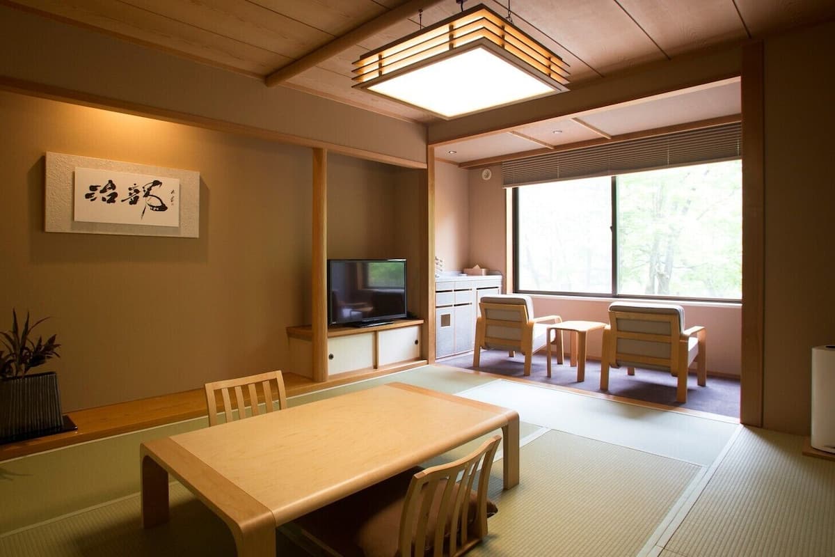 Una habitación japonesa tradicional con suelo de tatami, una mesa baja, sillas, un televisor y un ventanal grande.