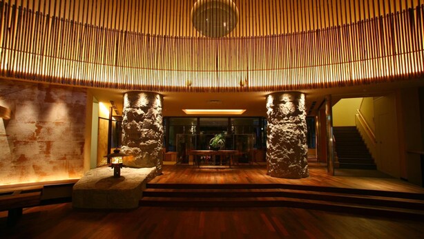 Eine Lobby mit Holzboden, Steinsäulen, einem zentralen Tisch mit Pflanze und einer Treppe auf der rechten Seite.