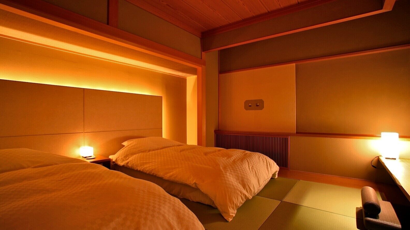 A hotel room with two beds, a bedside table, a nightstand, a wall-mounted heater, and a traditional Japanese tatami mat floor.