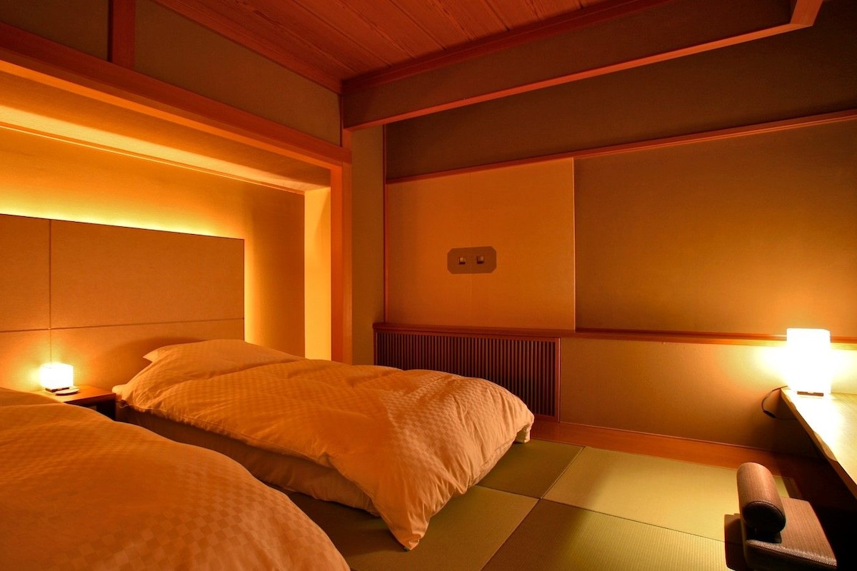 A hotel room with two beds, a bedside table, a nightstand, a wall-mounted heater, and a traditional Japanese tatami mat floor.