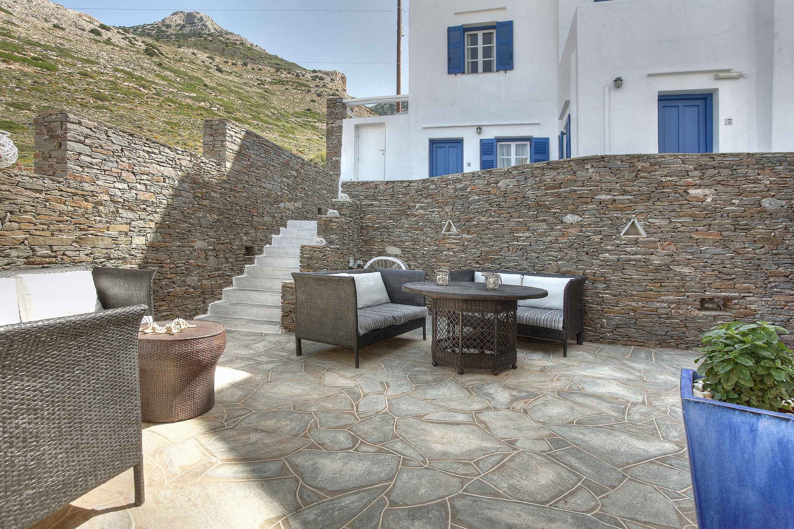 A stone-floored patio with wicker furniture and a table, overlooking a hillside.