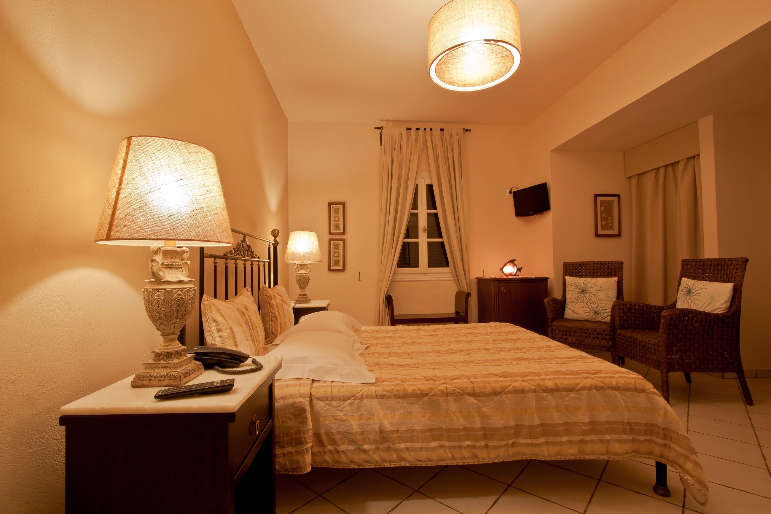 Une chambre à coucher avec un lit, deux lampes de chevet, une table de chevet, une télécommande, une lampe décorative, une chaise et un téléviseur fixé au mur.