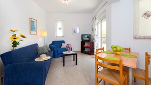 A living room with a blue sofa, a small table with a vase of flowers, a television, and a dining area with a table and chairs. - Hidden garden Rhodes Apartments (Rhodes)