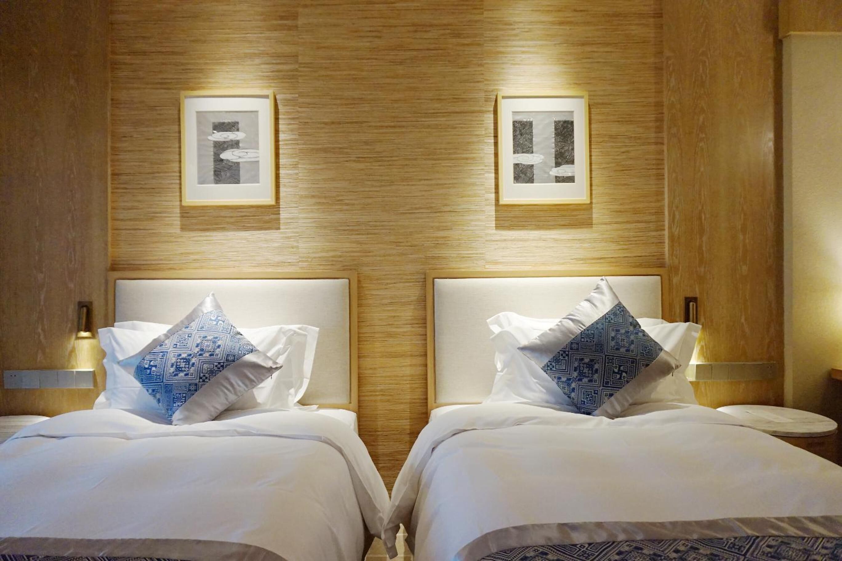 A hotel room with two beds, each with a headboard, white bedding, and patterned pillows. There are framed pictures on the wall above the beds and bedside lamps on either side.