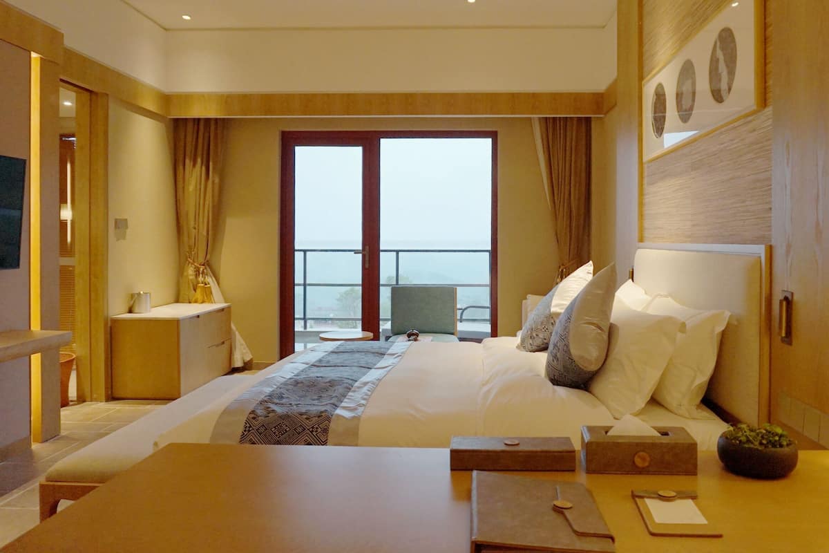 A hotel room with a large bed, a bedside table, a TV, and a balcony with a view.
