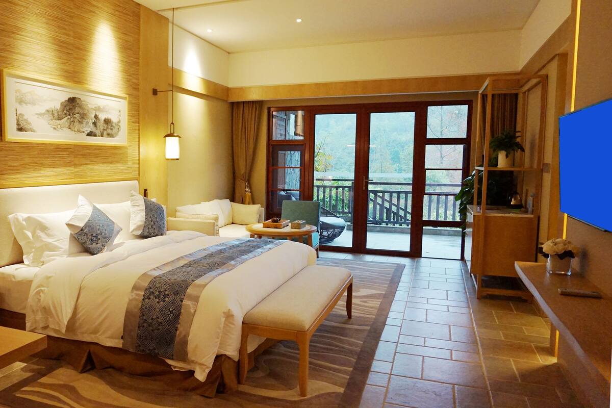 A hotel room with a large bed, a desk, a chair, a TV, and a balcony with a view.