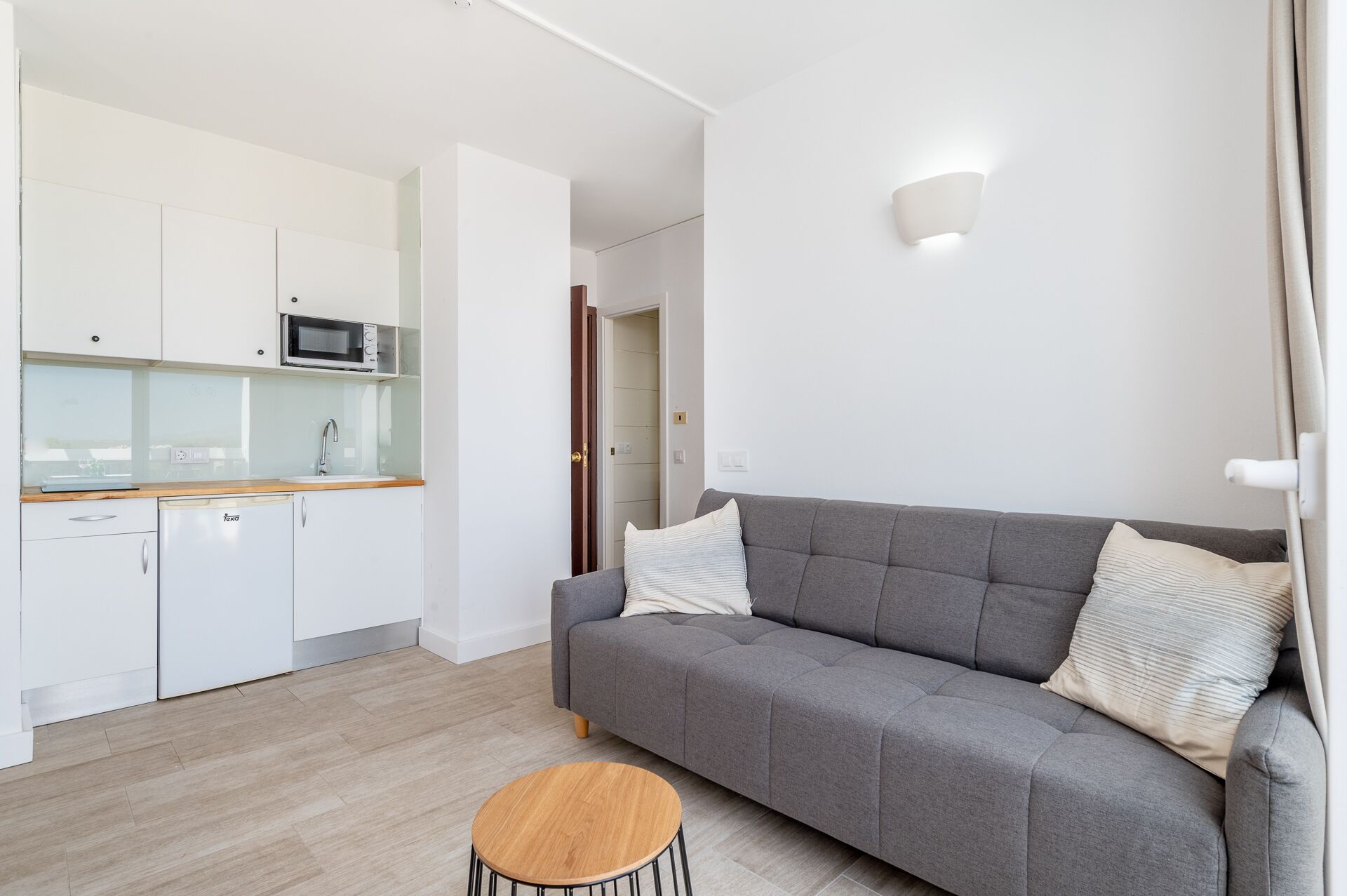 Un appartement moderne avec un canapé gris, une table basse en bois et un coin cuisine doté de meubles blancs et d’un four à micro-ondes.