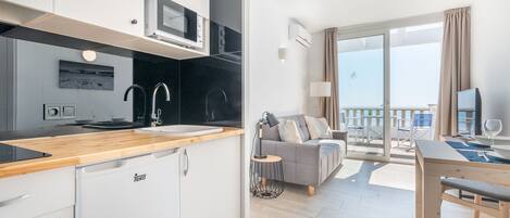 A modern kitchen with a microwave, sink, and white cabinets. There is a dining area with a table and chairs, and a balcony with a view of the sea.