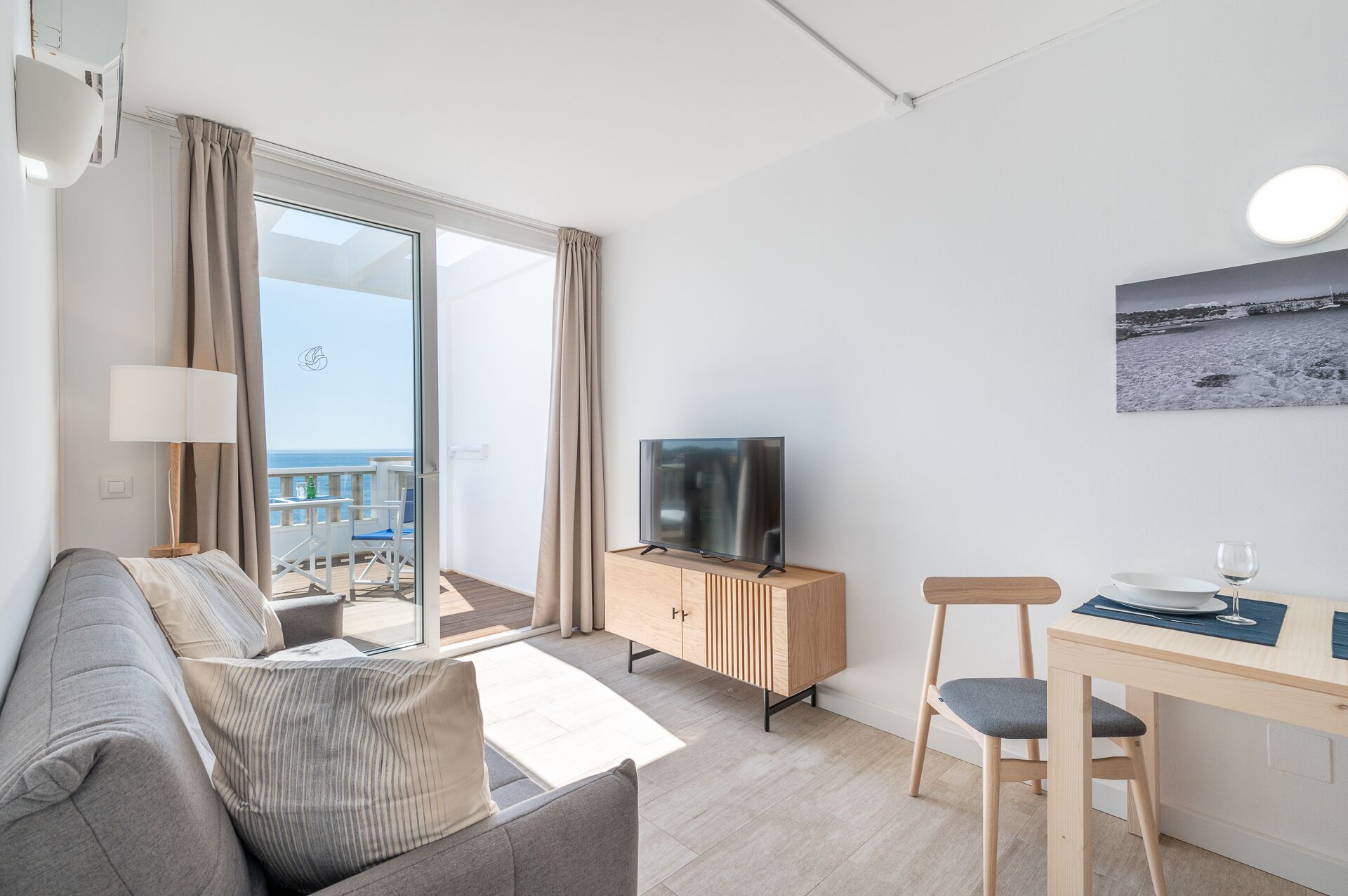 A modern living room with a sofa, a TV on a wooden cabinet, a dining area with a table and chair, and a balcony with a view of the sea.