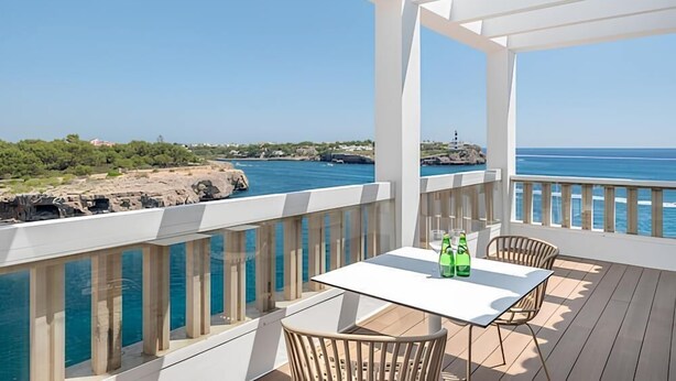 Un balcon avec une table et des chaises blanches, donnant sur une zone côtière avec un phare et un terrain rocheux.