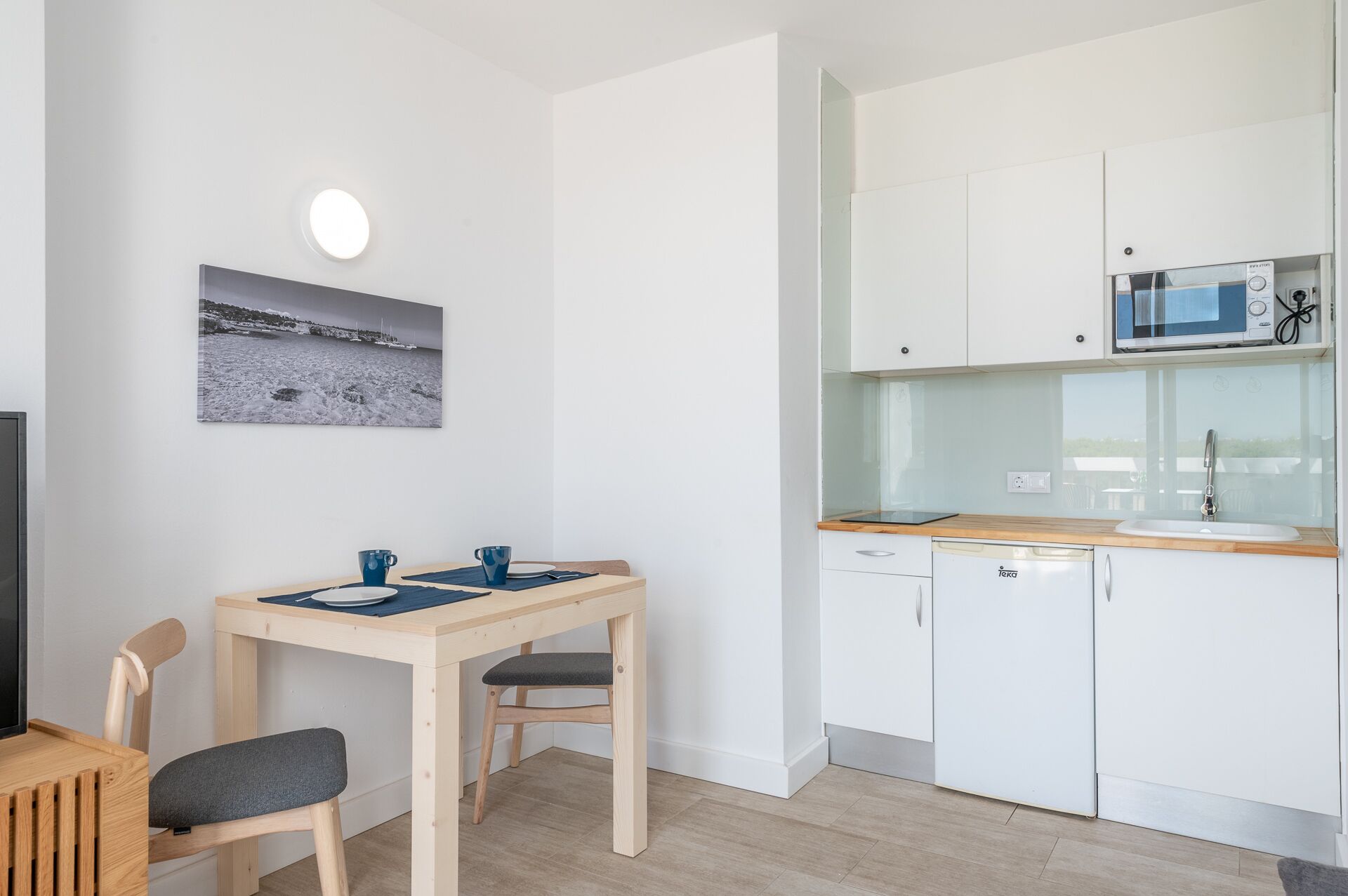 Une cuisine moderne avec des armoires blanches, un four à micro-ondes et un lave-vaisselle. On y trouve une table à manger avec deux chaises et, au mur, un tableau représentant un paysage enneigé.