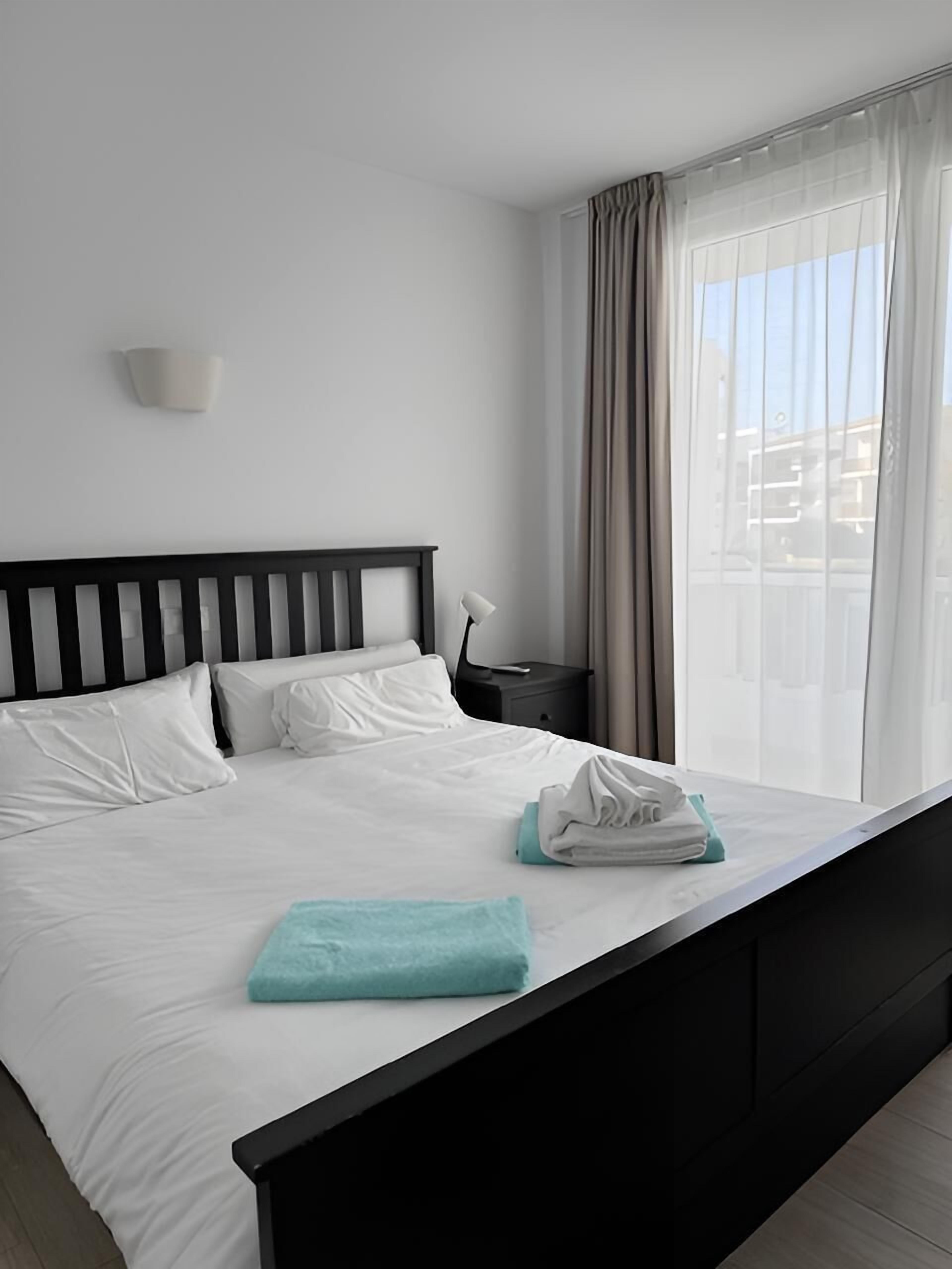 Habitación de hotel moderna con una cama grande, ropa de cama blanca, un cabecero negro, una mesita de noche, una lámpara y vistas de la ciudad a través de cortinas translúcidas.