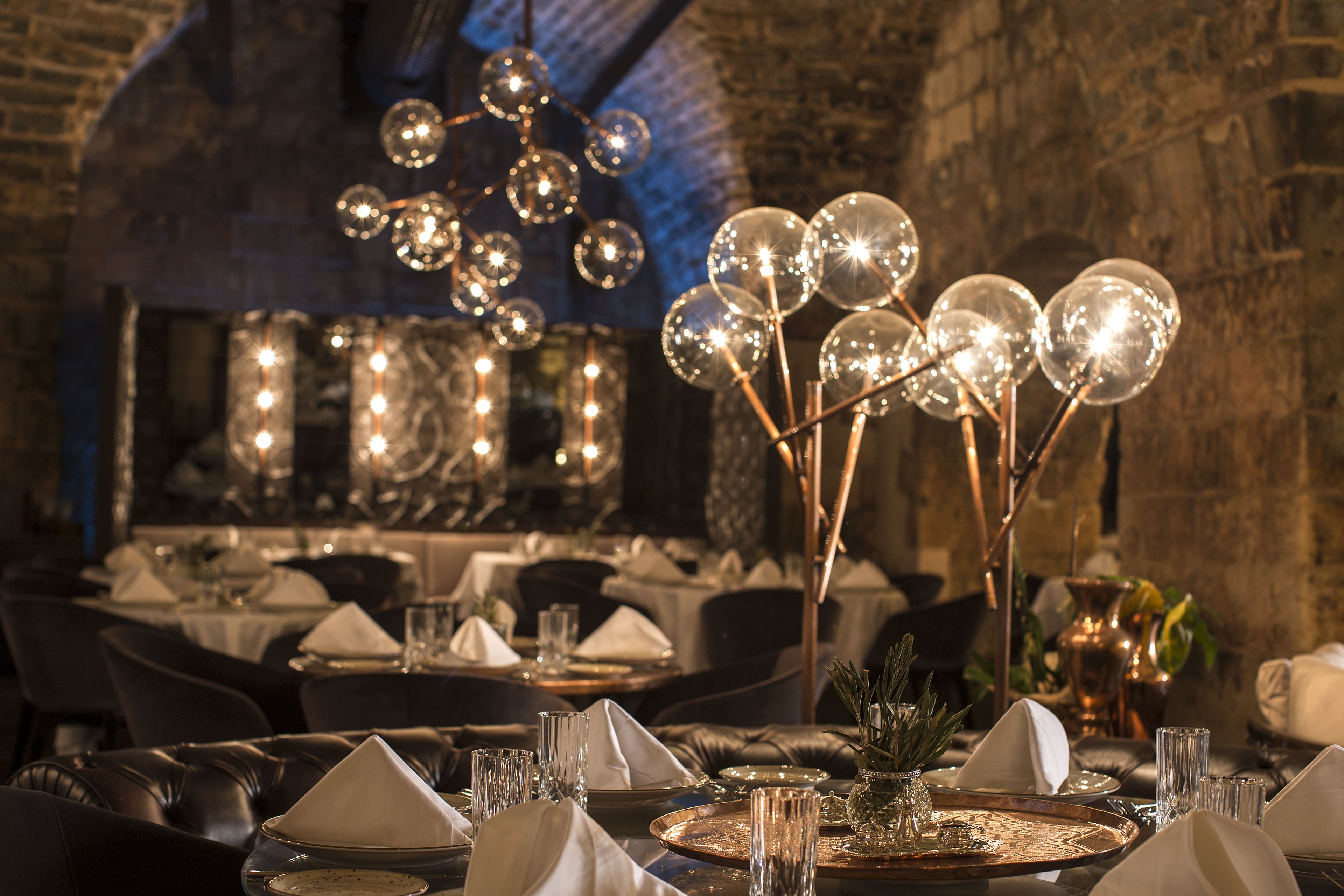 A restaurant setting with tables set for dining, featuring elegant table settings, clear glassware, and a decorative light fixture.