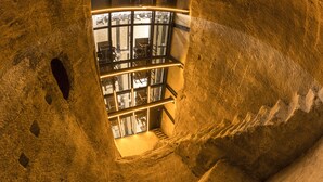 Un escalier en colimaçon, situé dans une structure ressemblant à une grotte, offre une vue sur une pièce dotée de fenêtres et d’un autre escalier menant vers le bas.