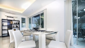 A modern dining area with a glass table and white chairs, large windows, and a view of the outdoors. - Villa Cheloni 3 Bedrooms (Kamala)